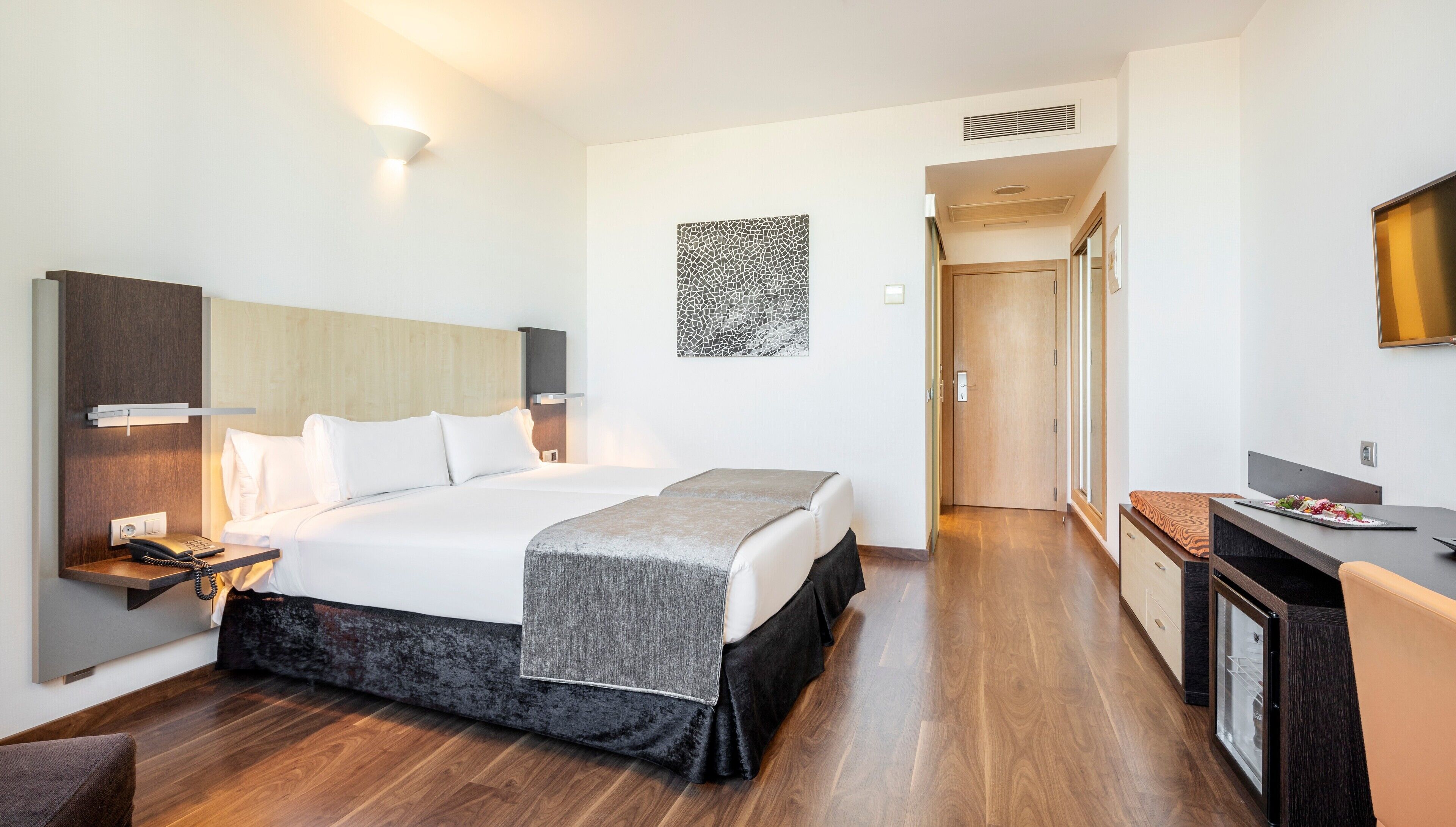A modern hotel room with a large bed, bedside table with a phone, a desk with a computer, and a small refrigerator.