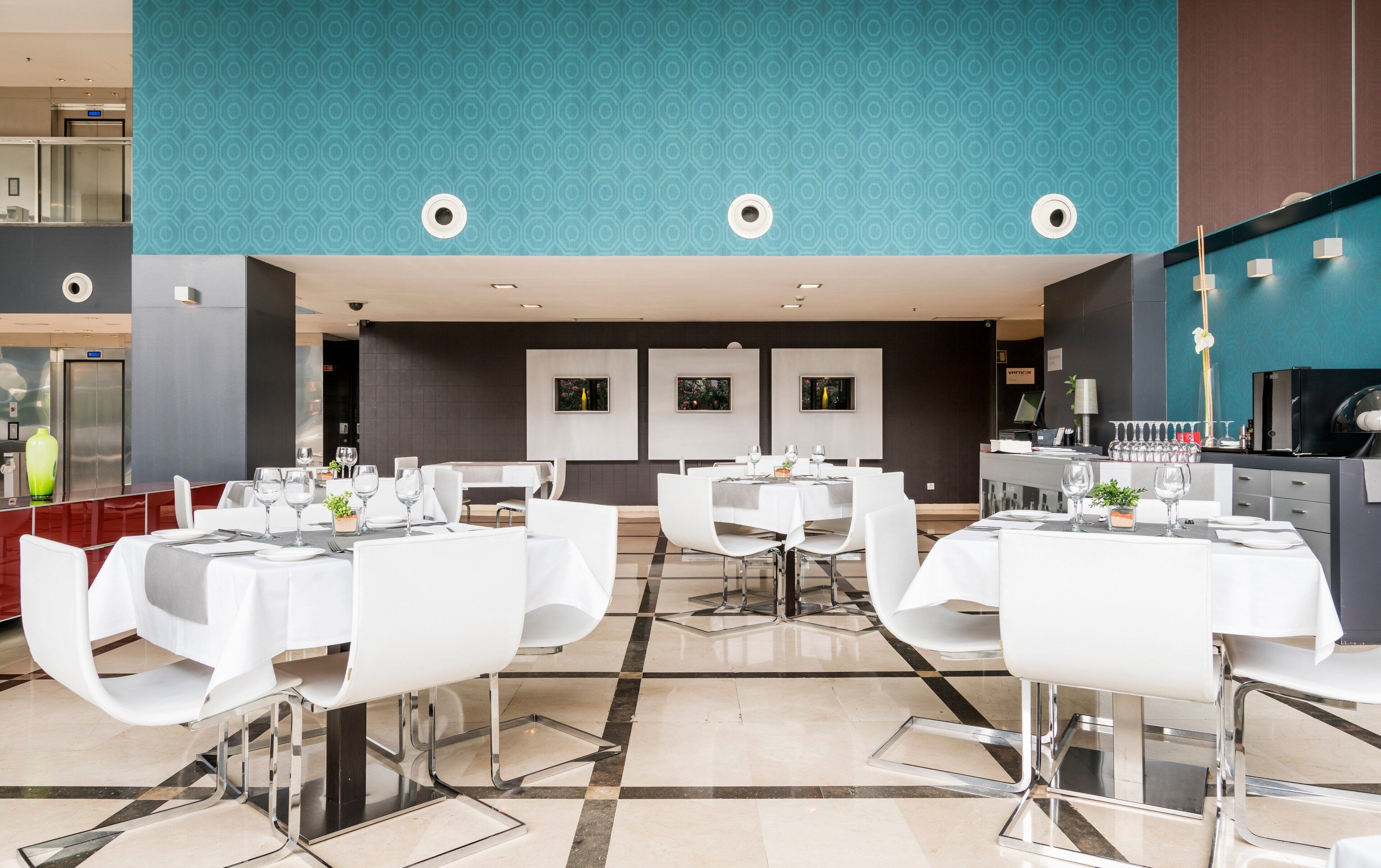 A modern dining area with white tables and chairs, a patterned ceiling, and a kitchen area with a black refrigerator.