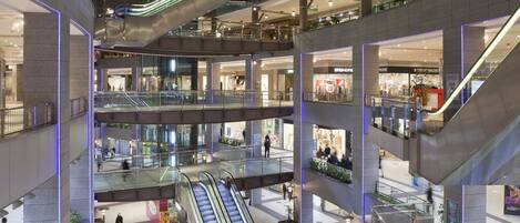 A multi-level shopping mall with escalators connecting floors, glass railings, and potted plants.