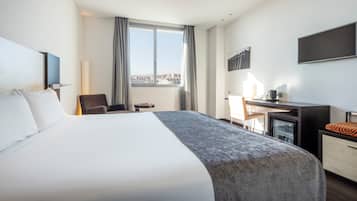 A hotel room with a large bed, a desk, a chair, a TV, and a city view.