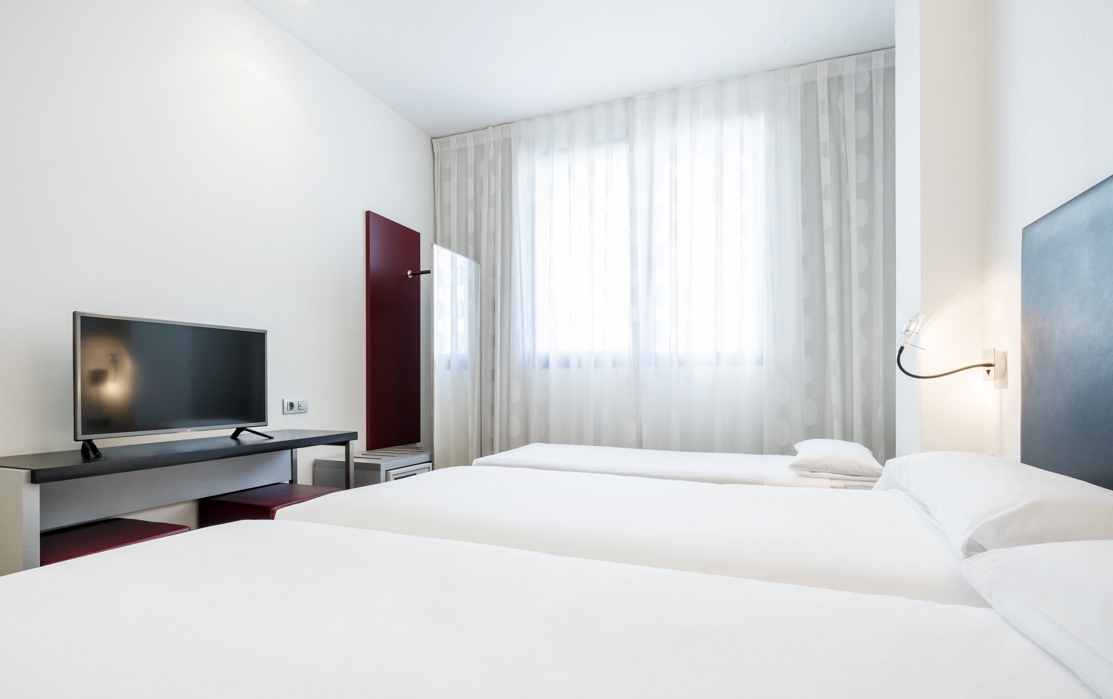 Una habitación de hotel con cama, escritorio con televisor, ventana con cortinas translúcidas y lámpara fijada a la pared.