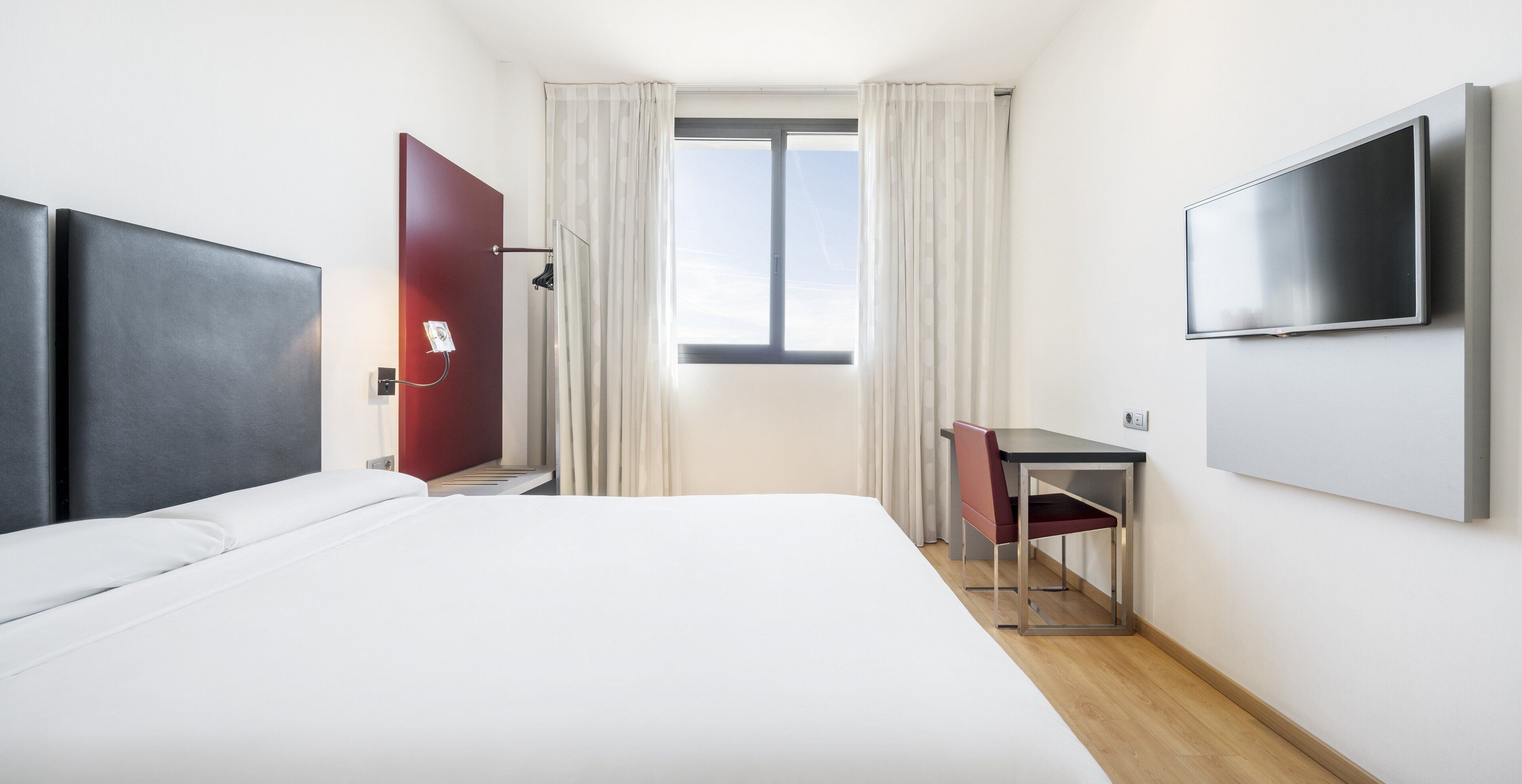 Una camera d'albergo con un letto, una scrivania, una sedia, una TV e una finestra con tende.