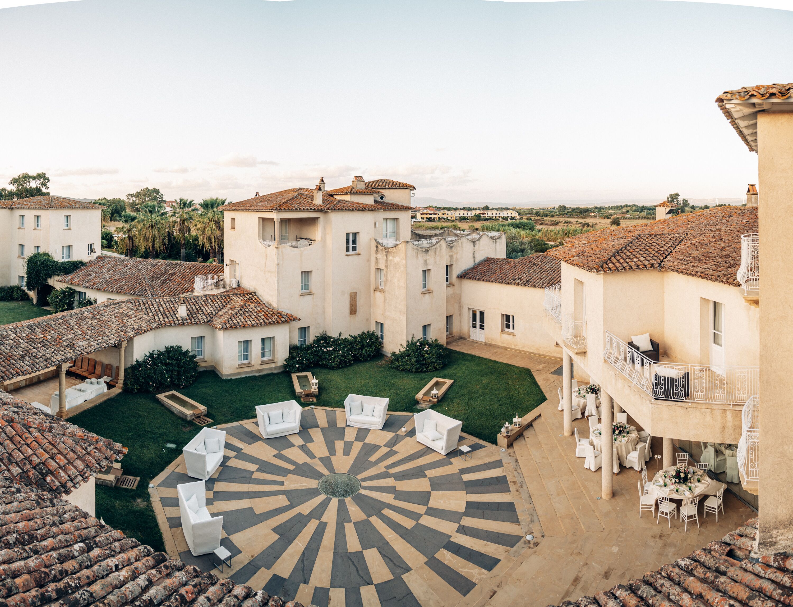 Un patio con un área de estar circular en el centro, rodeado de edificios con tejados de teja y una piscina.