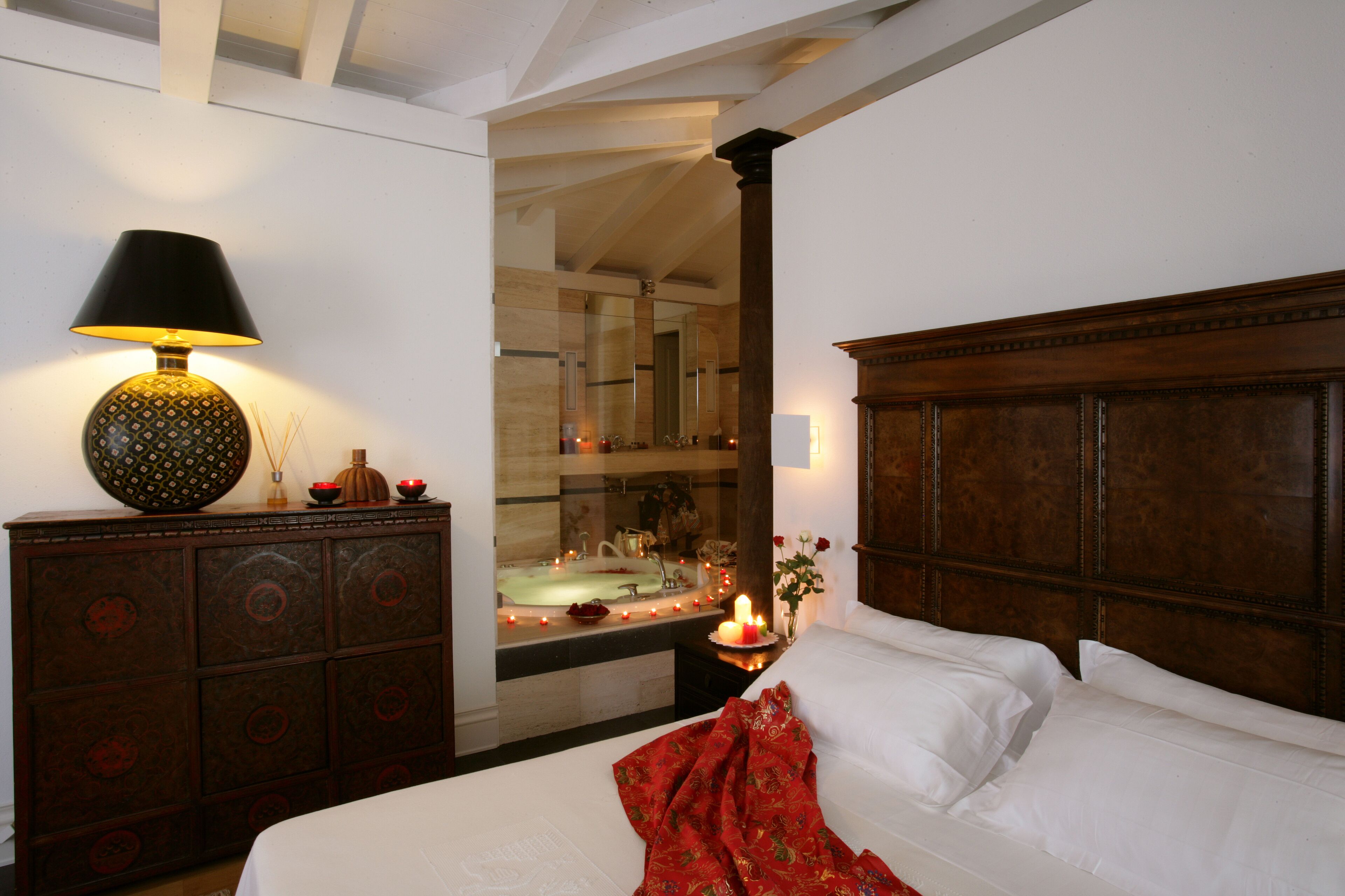Una camera da letto con una testiera in legno, un comodino con una lampada, uno specchio e vista sul bagno con una vasca.