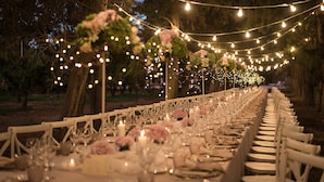 Una larga mesa de comedor dispuesta para un evento formal, rodeada de sillas blancas, con luces de cuerda en el techo y centros de mesa florales.