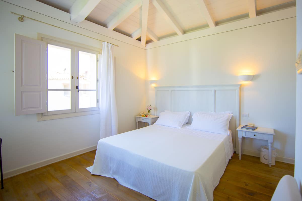 A bedroom with a large bed, two bedside tables, a nightstand, a window with white shutters, and a wooden ceiling.