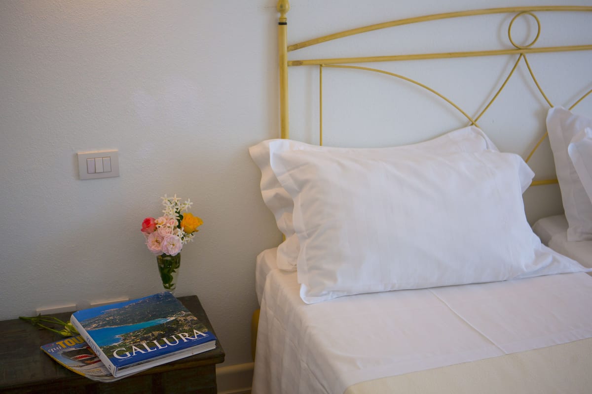 A neatly made bed with white bedding, a small bedside table with a book and a vase of flowers, and a simple wall switch.