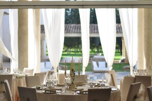 Un ambiente formal para comer al aire libre, con mesas dispuestas para una comida, manteles blancos, una elegante vajilla y vistas a un jardín y un edificio al fondo.