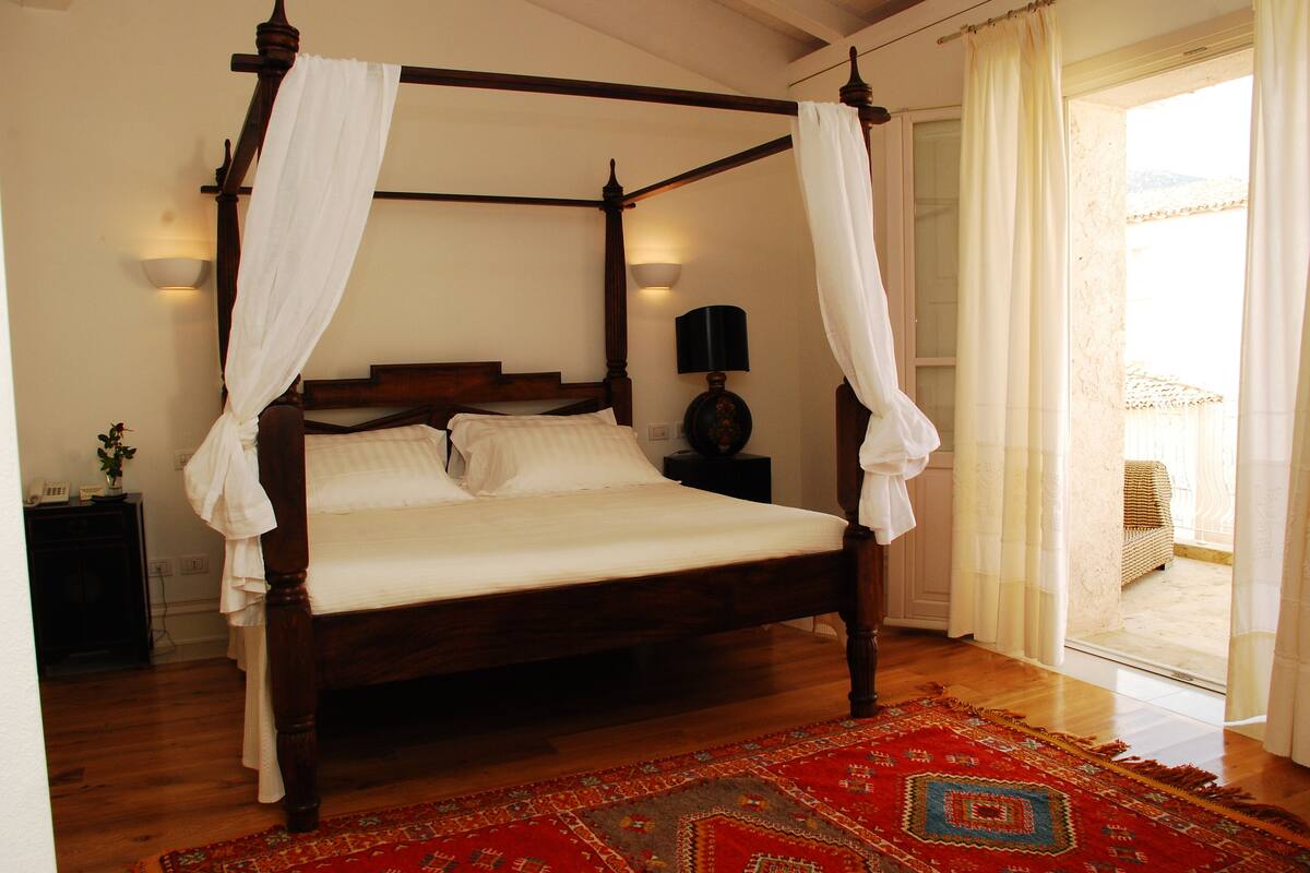 A four-poster bed with white linens, a wooden headboard, and a red patterned rug.