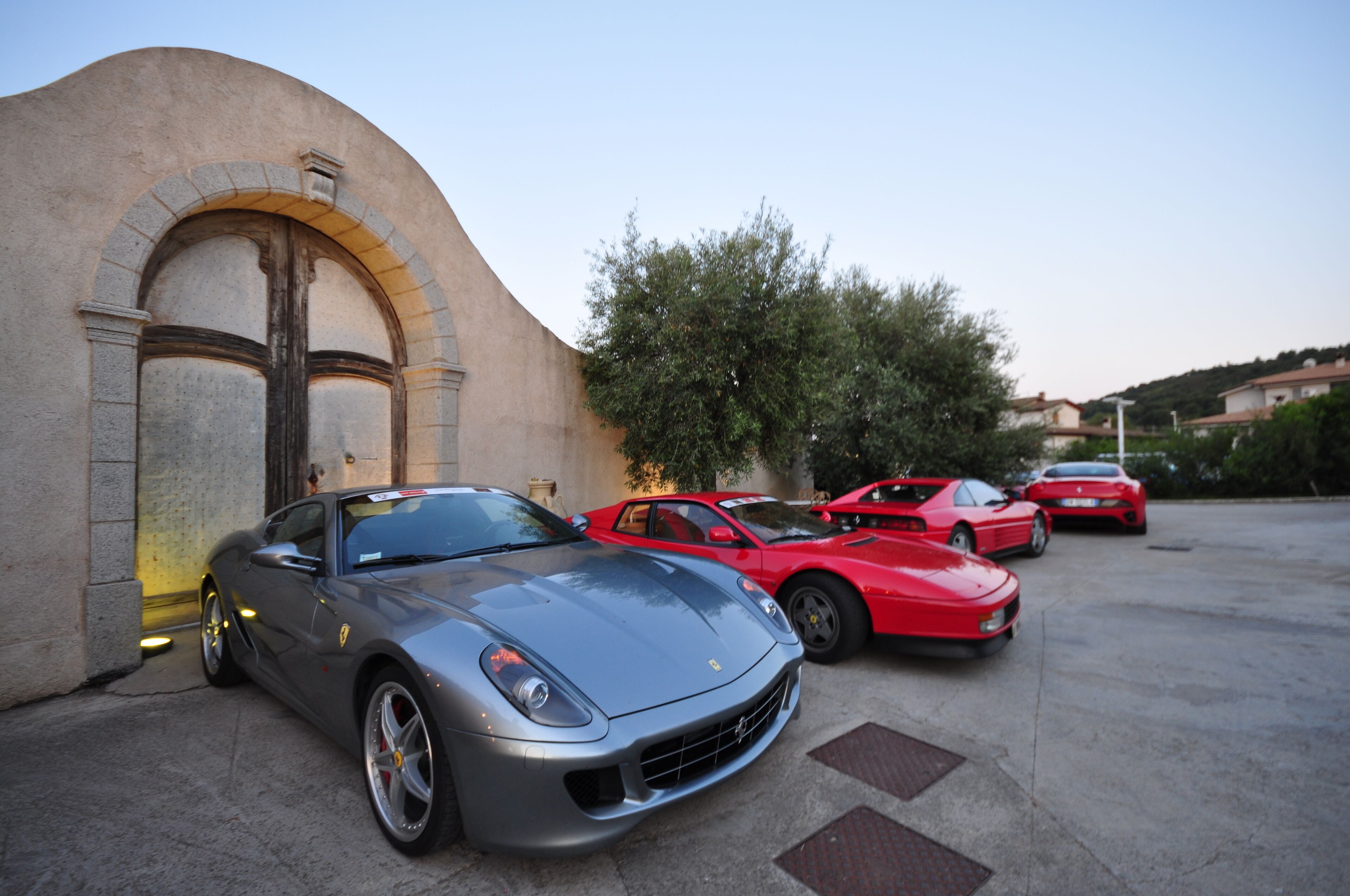 A parking area with several sports cars, including a grey Ferrari and red sports cars, parked near a building with a curved facade and large doors.