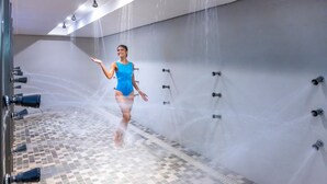 A woman in a blue swimsuit in a shower area with multiple water jets, tiled floor, and a wall-mounted showerhead. - Serhs Natal Grand Hotel & Resort (Natal)