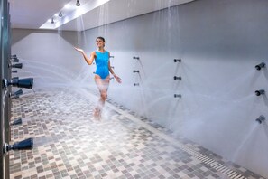 A woman in a blue swimsuit in a shower area with multiple water jets, tiled floor, and a wall-mounted showerhead. - Serhs Natal Grand Hotel & Resort (Natal)