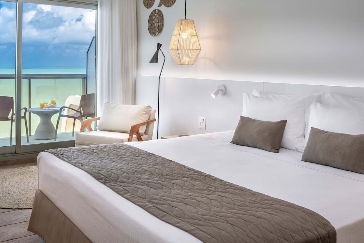A hotel room with a large bed, a view of the ocean, a balcony with chairs, and a table with drinks.
