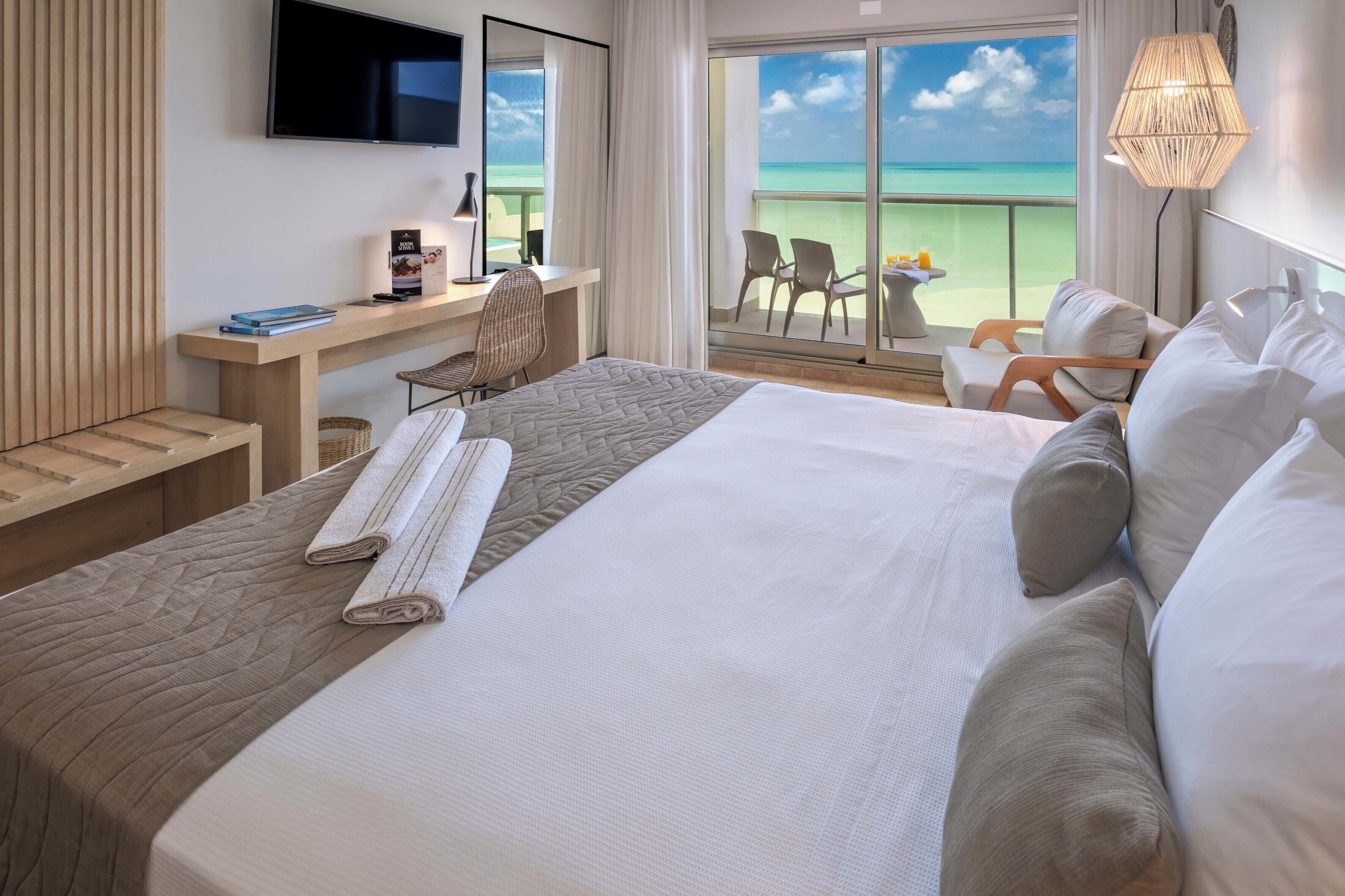 A hotel room with a large bed, a desk, a chair, a TV, and a balcony with a view of the ocean.