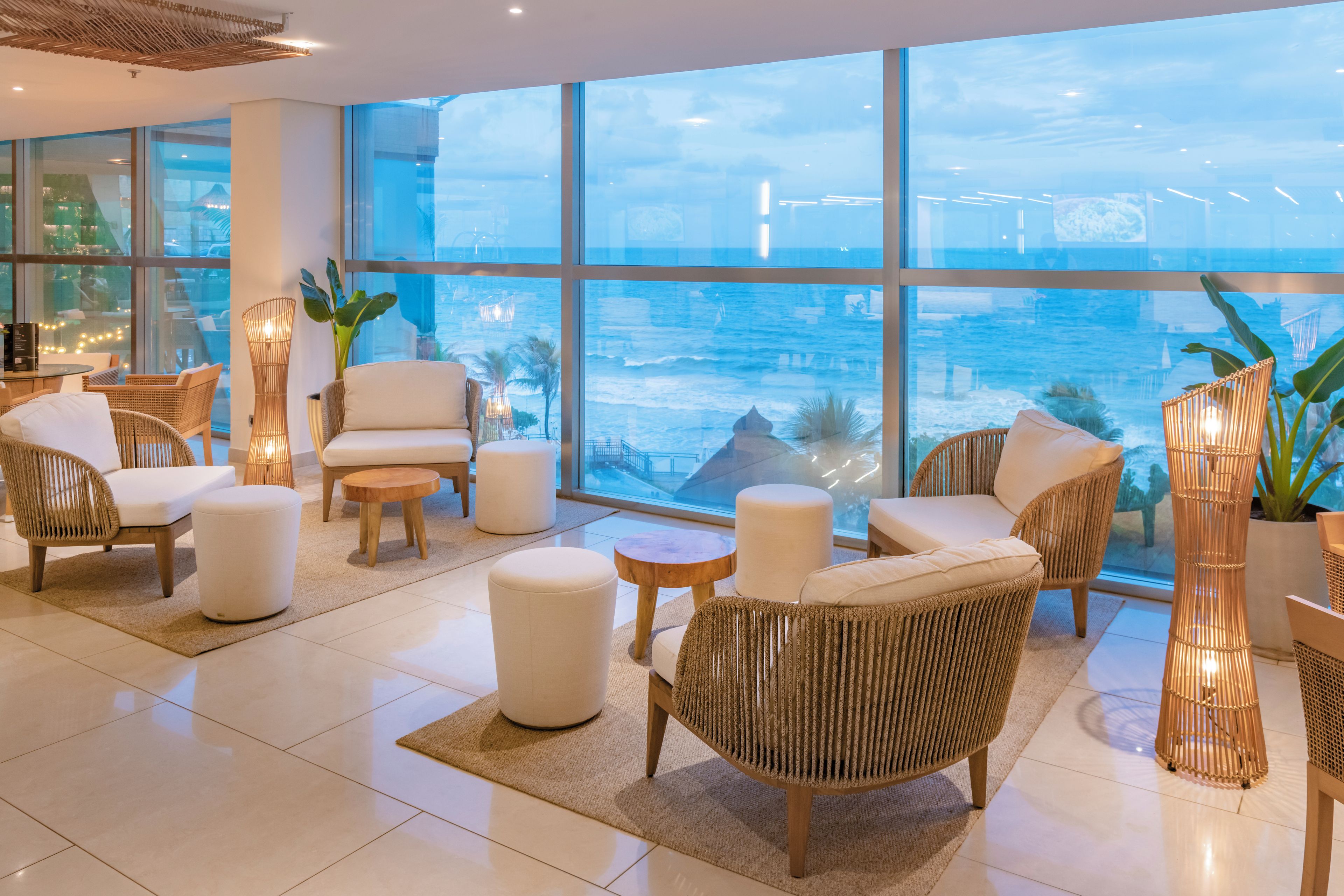 A modern lounge area with wicker furniture, white cushions, and large windows offering a view of the sea.