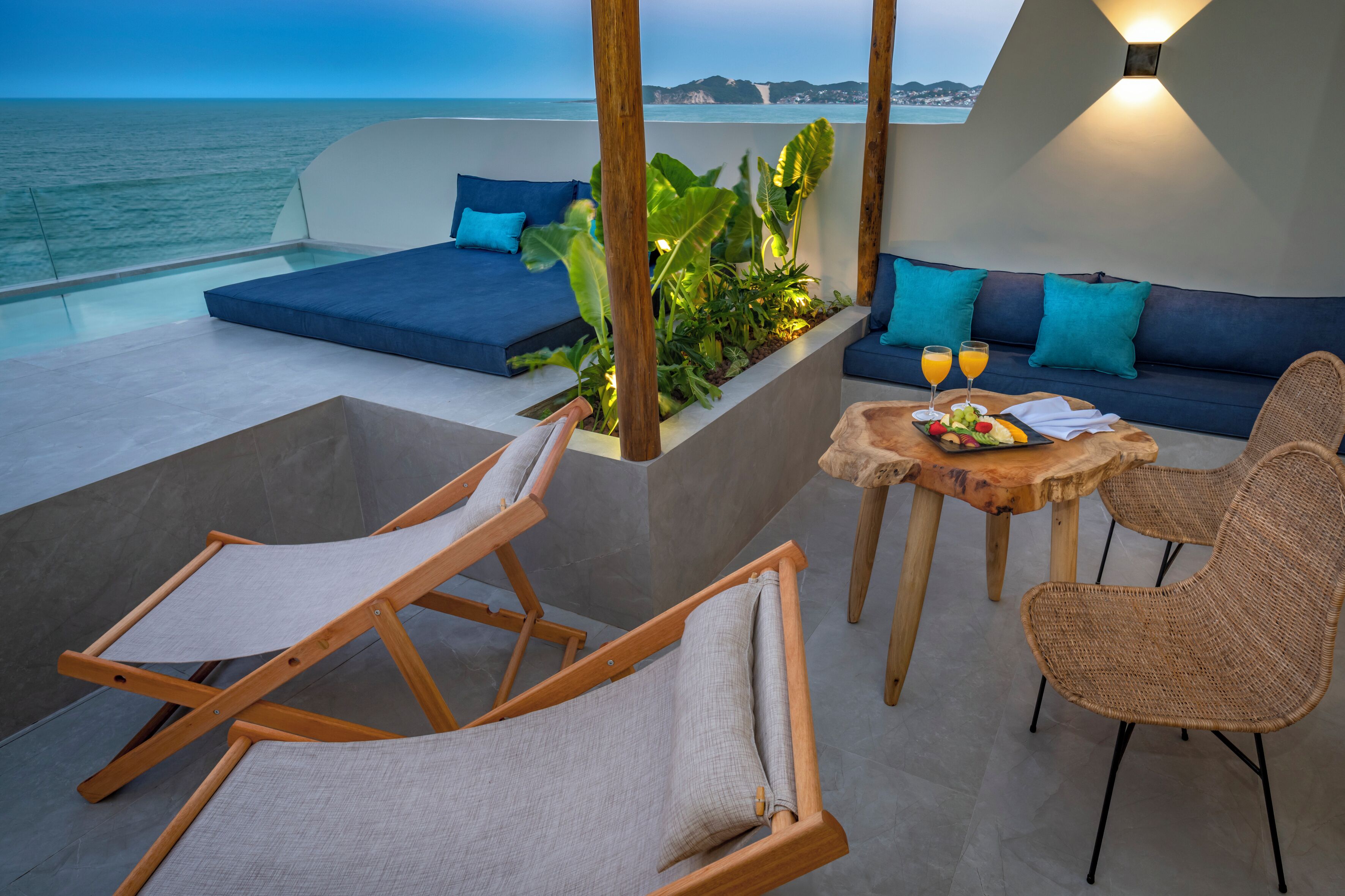 Um terraço com vista para o mar, um sofá, uma mesa com frutas e bebidas, e cadeiras de descanso.