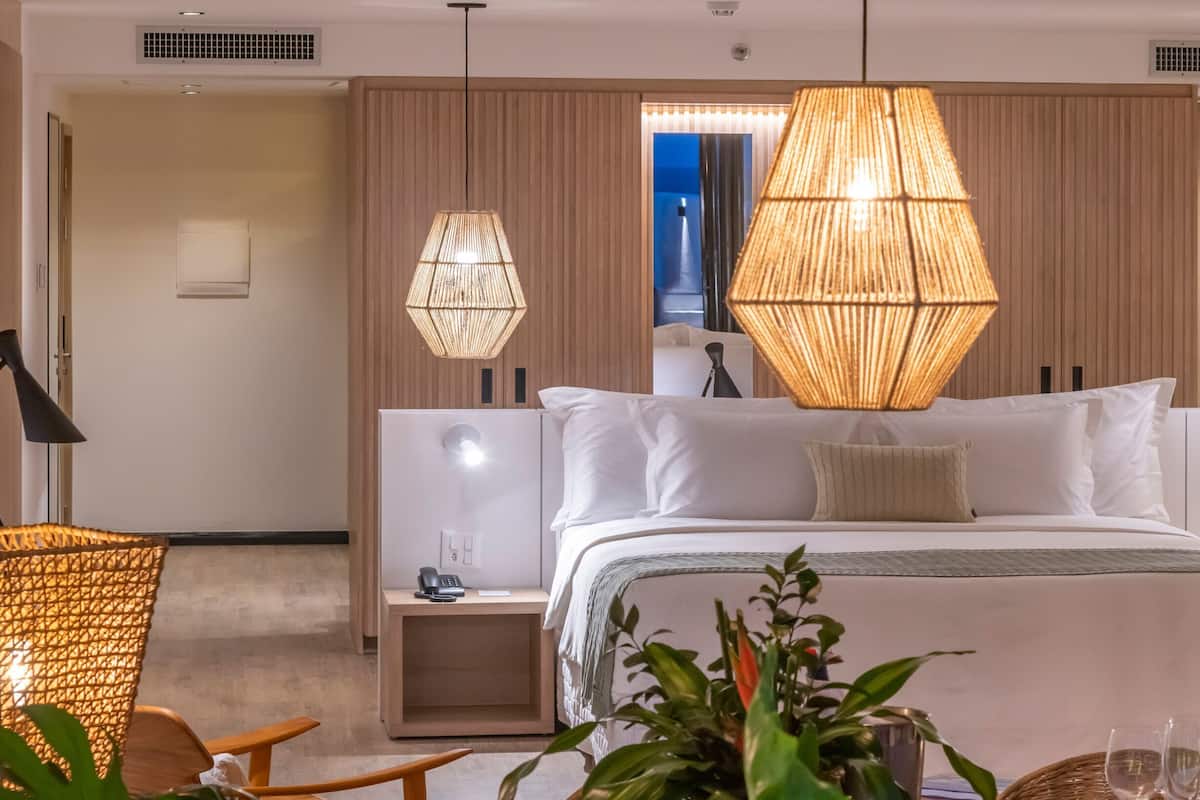 A modern hotel room with a large bed, two bedside lamps, a nightstand, a small table, and a potted plant.