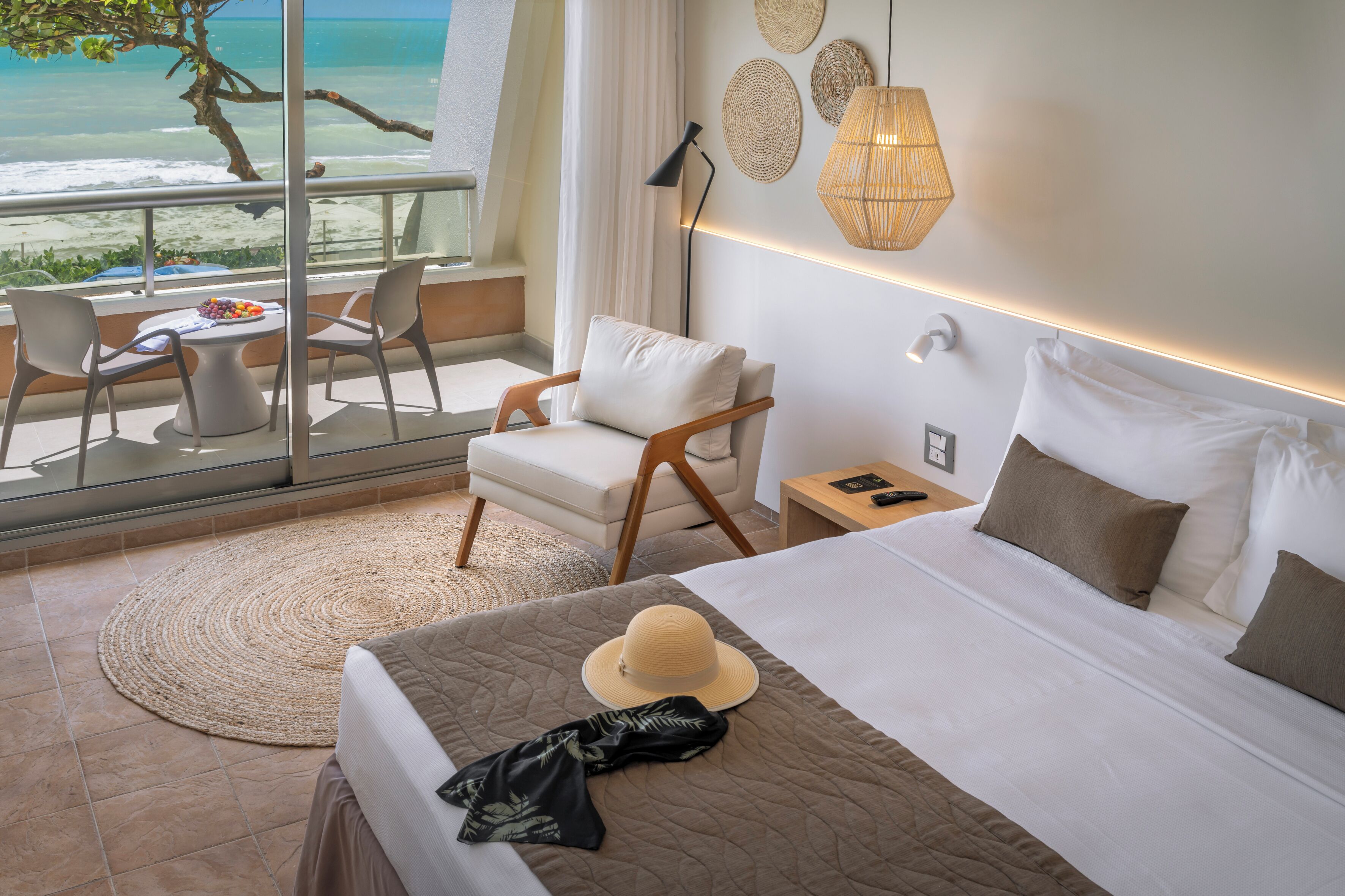Quarto de hotel com cama, uma cadeira, uma mesinha com frutas, vista para a praia e uma varanda com grade.