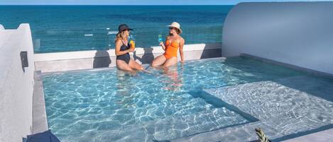 Zona de piscina con dos personas sentadas junto a la piscina, un frutero en el borde y vista despejada del océano.