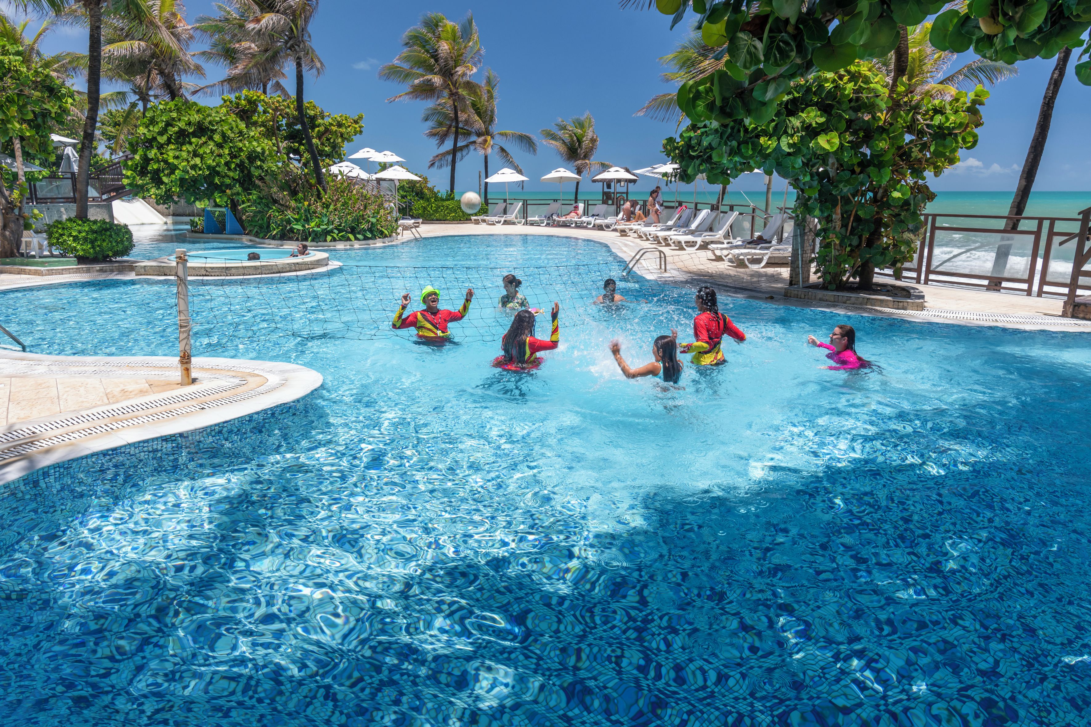 Una piscina con gente nadando, rodeada de palmeras y sombrillas de playa.