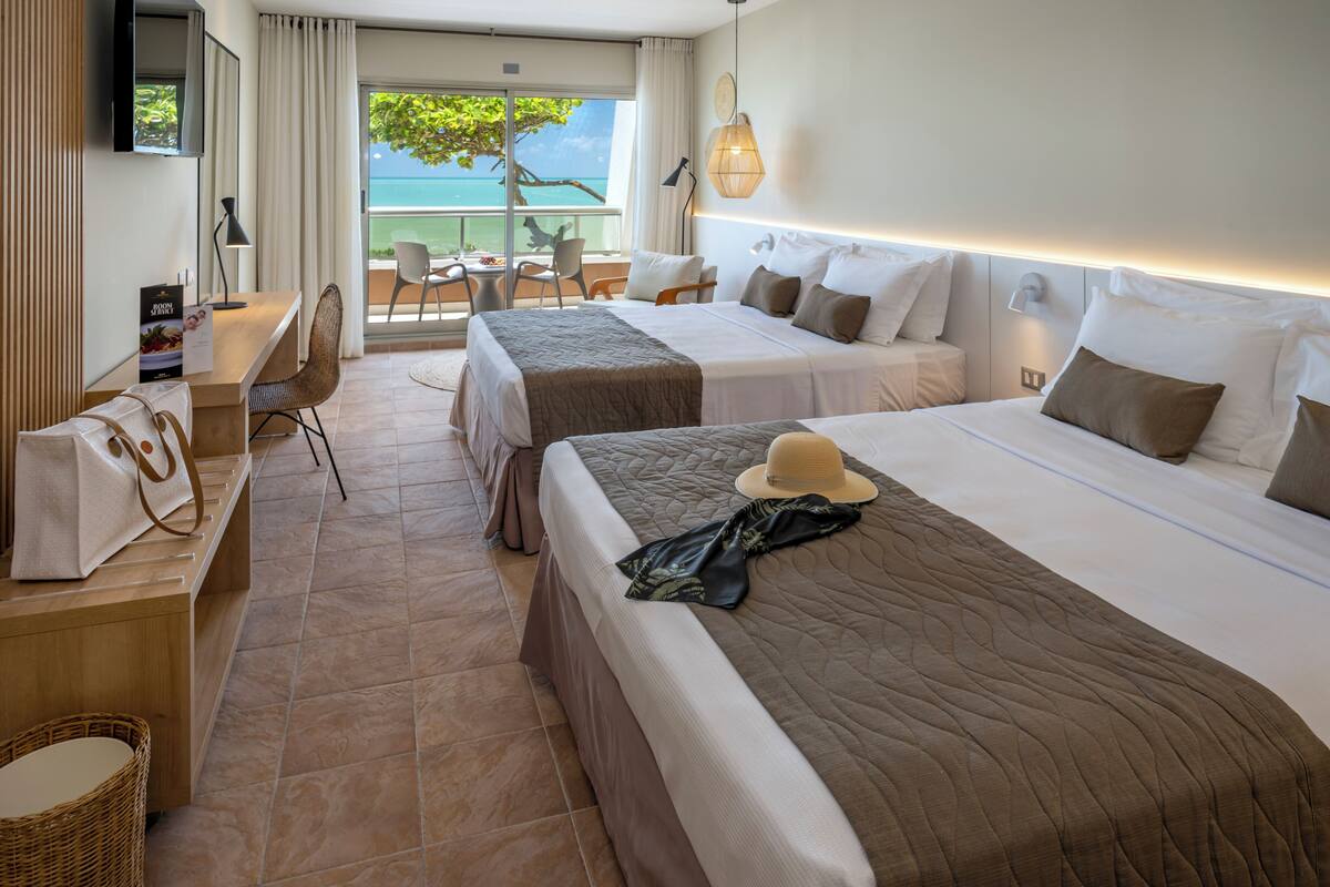 A hotel room with two beds, a desk, a chair, a TV, and a balcony with a view of the sea.