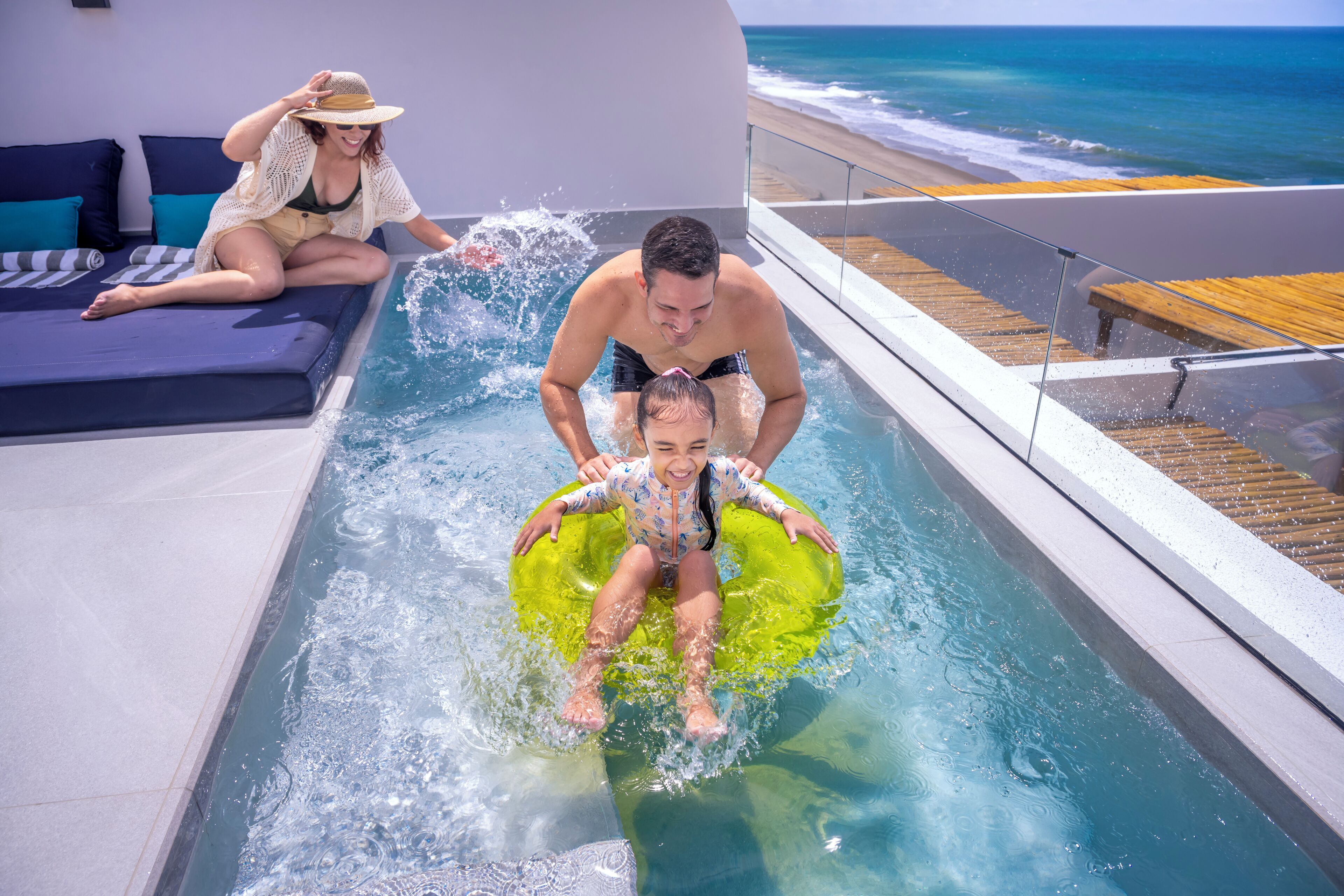 A pool area with a view of the beach, a lounge with blue cushions, and a person sitting on a lounge chair.