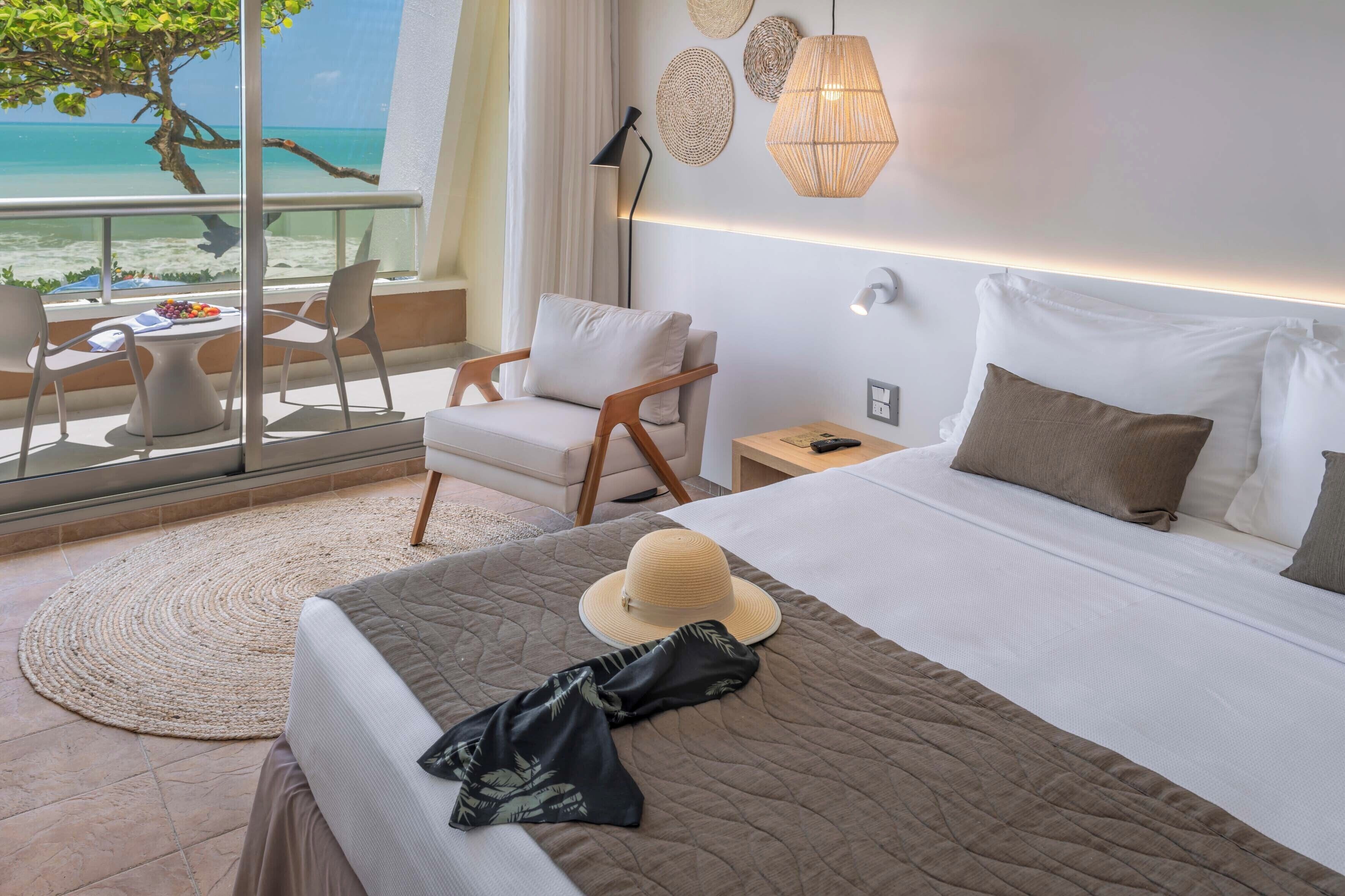 Habitación de hotel con una cama, vista a la playa, zona de comedor con sillas y una mesa con fruta.