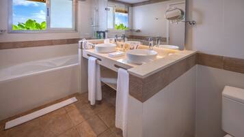 A modern bathroom with a bathtub, two sinks, a large mirror, and a window with a view of greenery.