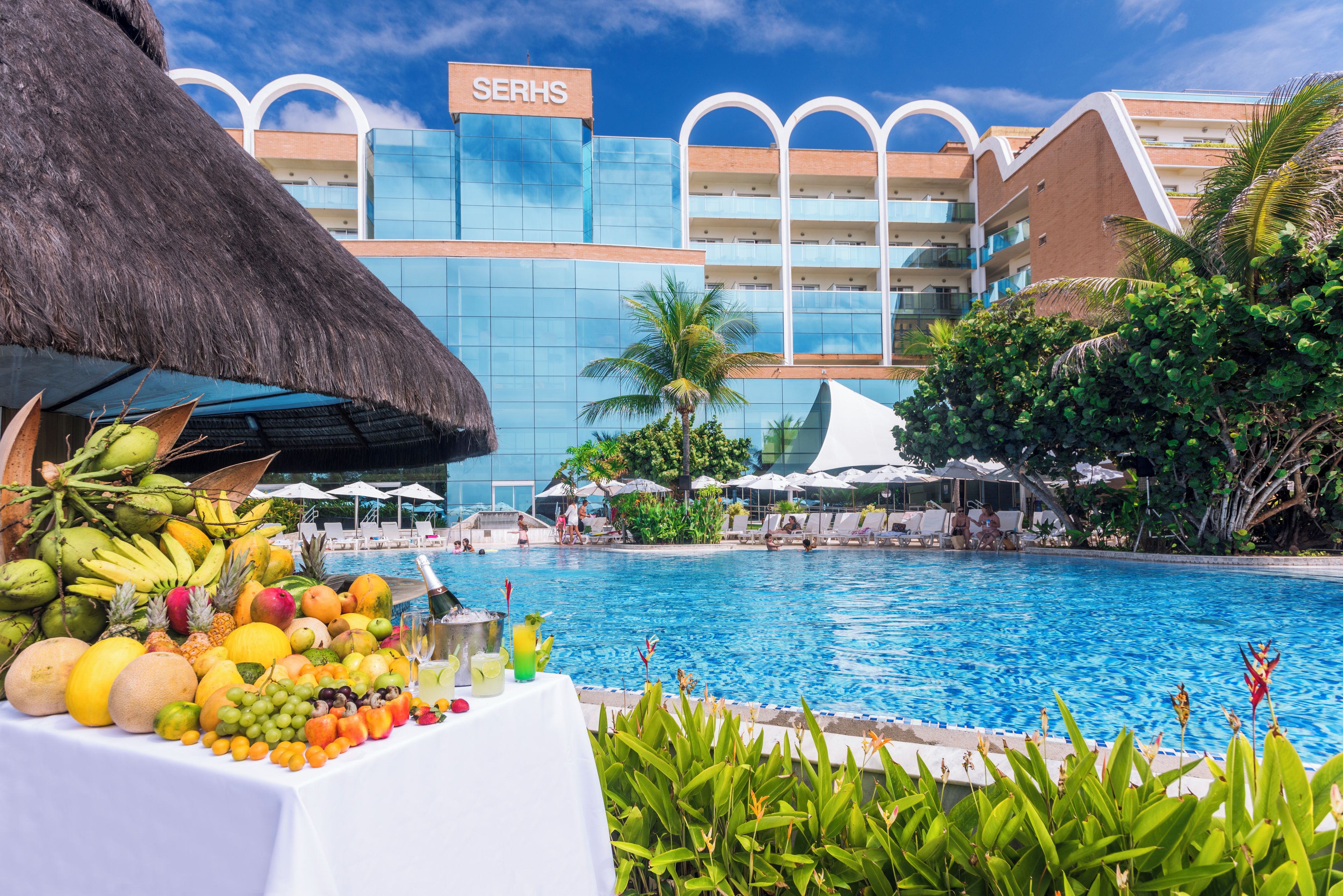 Un espace piscine agrémenté d’un présentoir de fruits et de boissons, d’un parasol en chaume, et un bâtiment d’hôtel avec des balcons en arrière-plan.