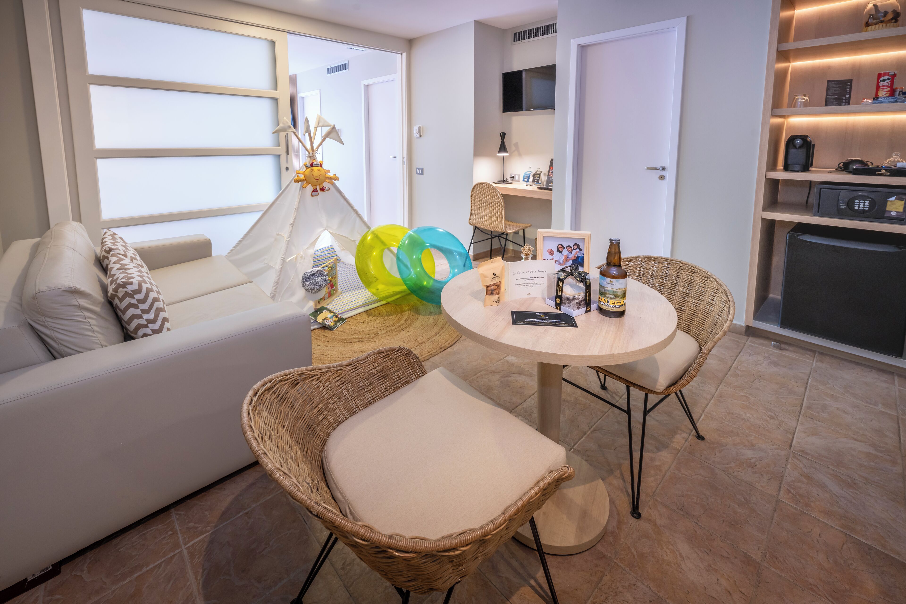Een moderne woonkamer met een bank, twee rieten fauteuils, een ronde tafel en een keukenhoek met een koelkast en planken.
