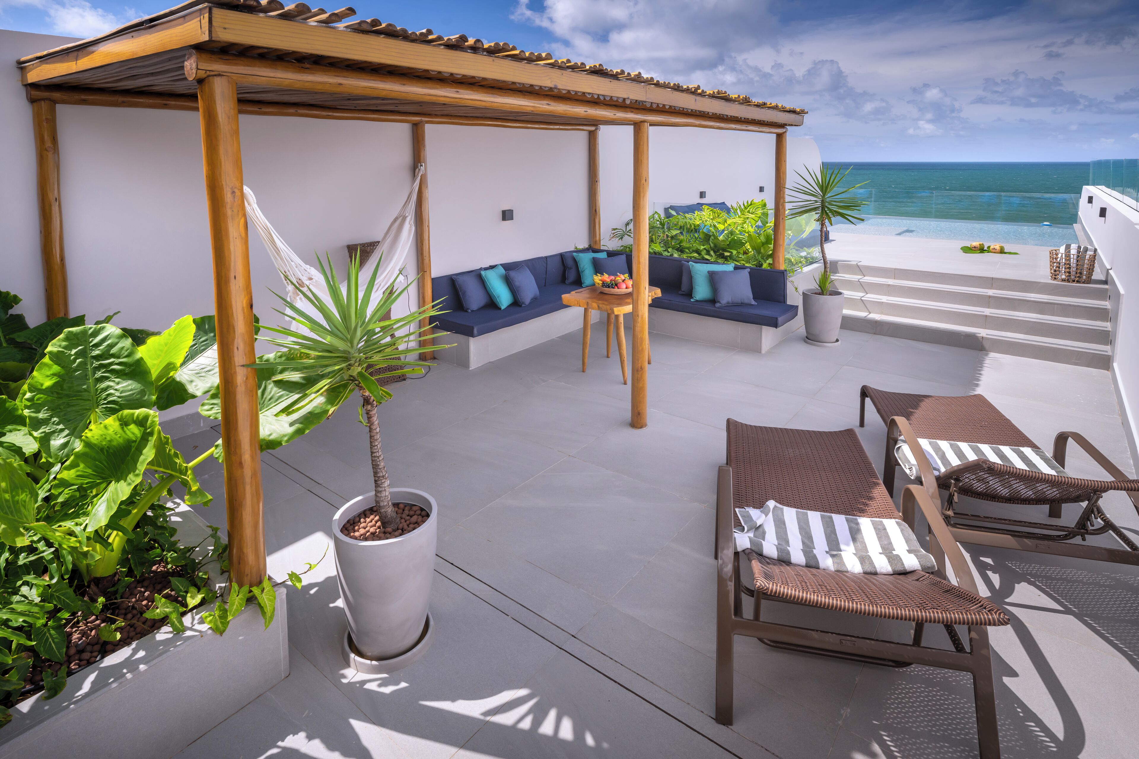 A rooftop terrace with a wooden pergola, a seating area with cushions, a small table with a fruit bowl, and lounge chairs.