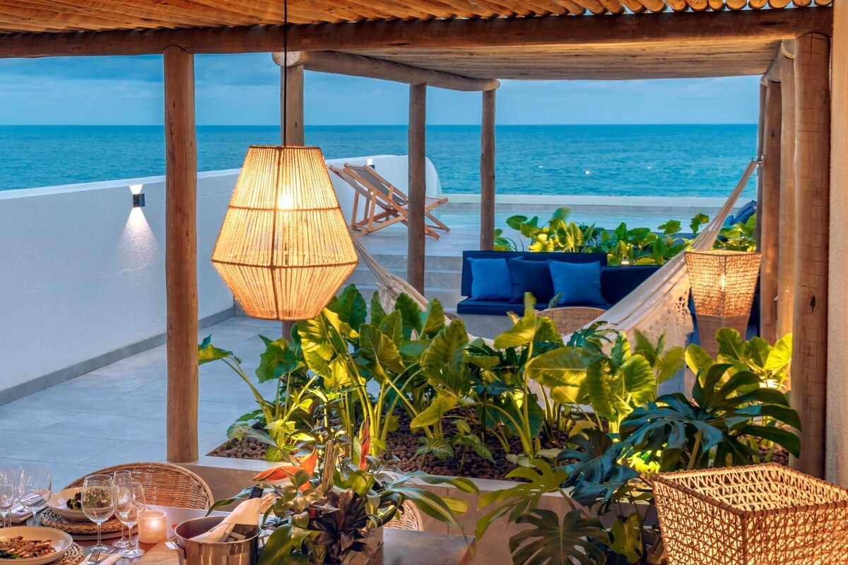A terrace with a view of the sea, wicker furniture, potted plants, and a hanging light fixture.