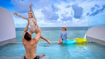 Cena à beira da piscina com um homem levantando uma criança, outra pessoa sentada com um boião, e vista clara do oceano sob um céu parcialmente nublado.