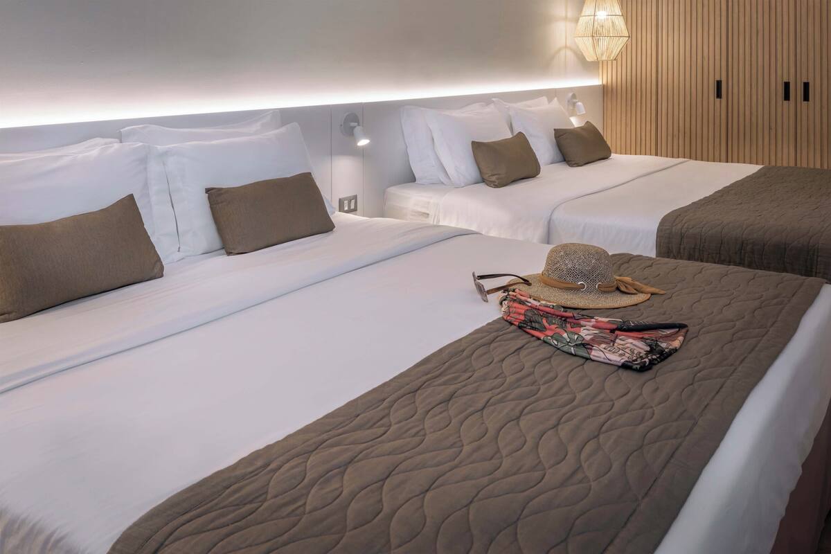 A hotel room with two beds, each with white linens and brown pillows. There is a woven hat and a patterned bag on one bed.