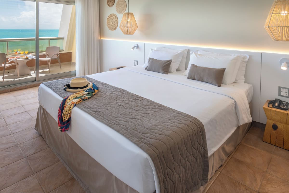 A hotel room with a large bed, a balcony with a view of the ocean, and a wooden nightstand with a phone.