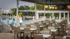 A covered outdoor seating area with wicker furniture and a pool in the background. - Antigoni Hotel (Paralimni)