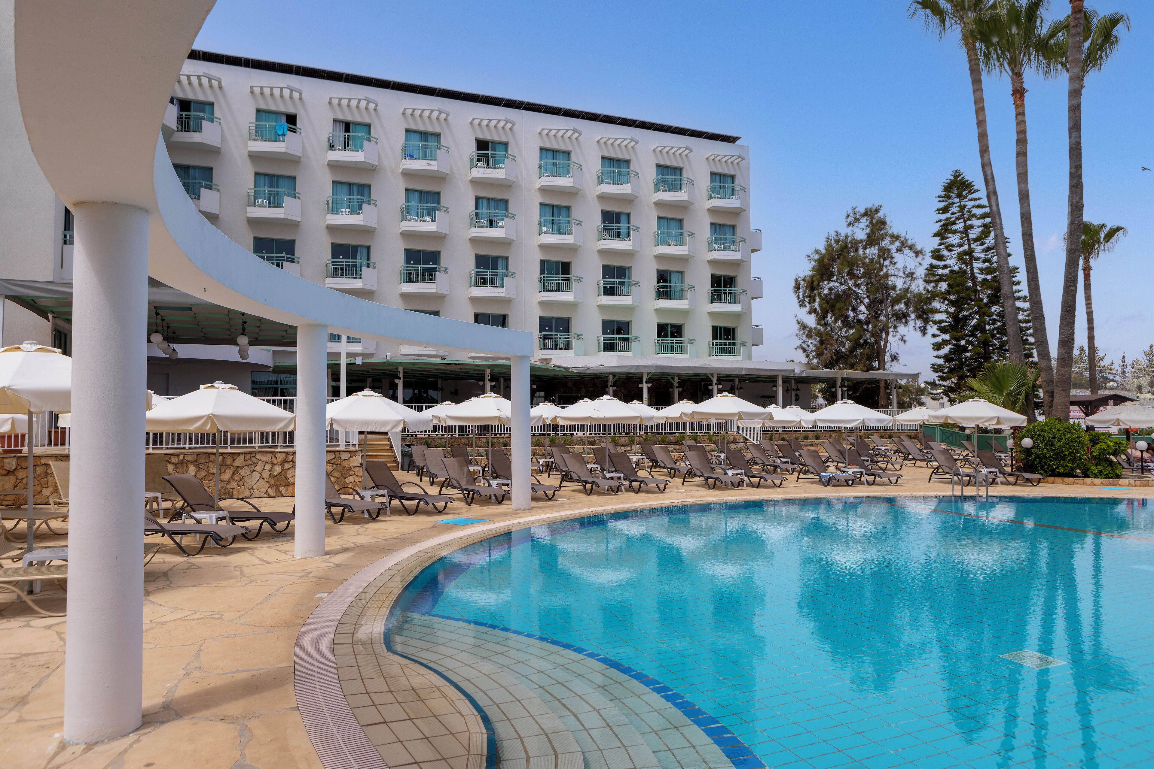 A swimming pool with lounge chairs and umbrellas, a building with balconies, and palm trees.