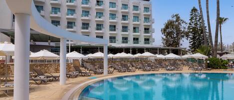 A swimming pool with lounge chairs and umbrellas, a building with balconies, and palm trees.