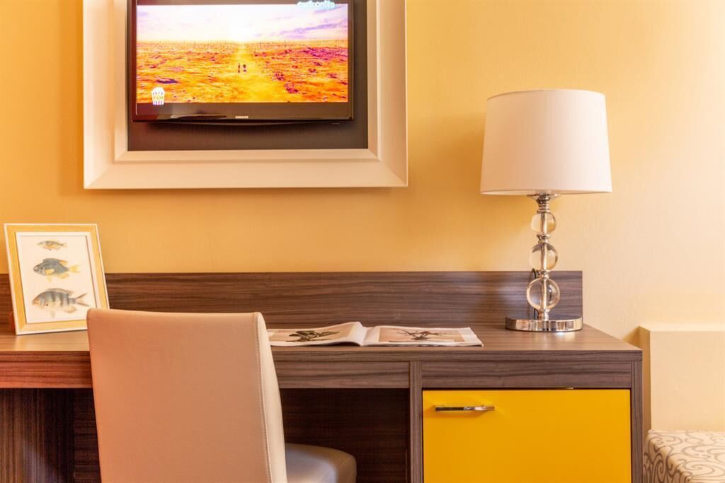 Habitación de hotel con una televisión montada en la pared, una mesita de noche con lámpara, una silla y un armario amarillo.