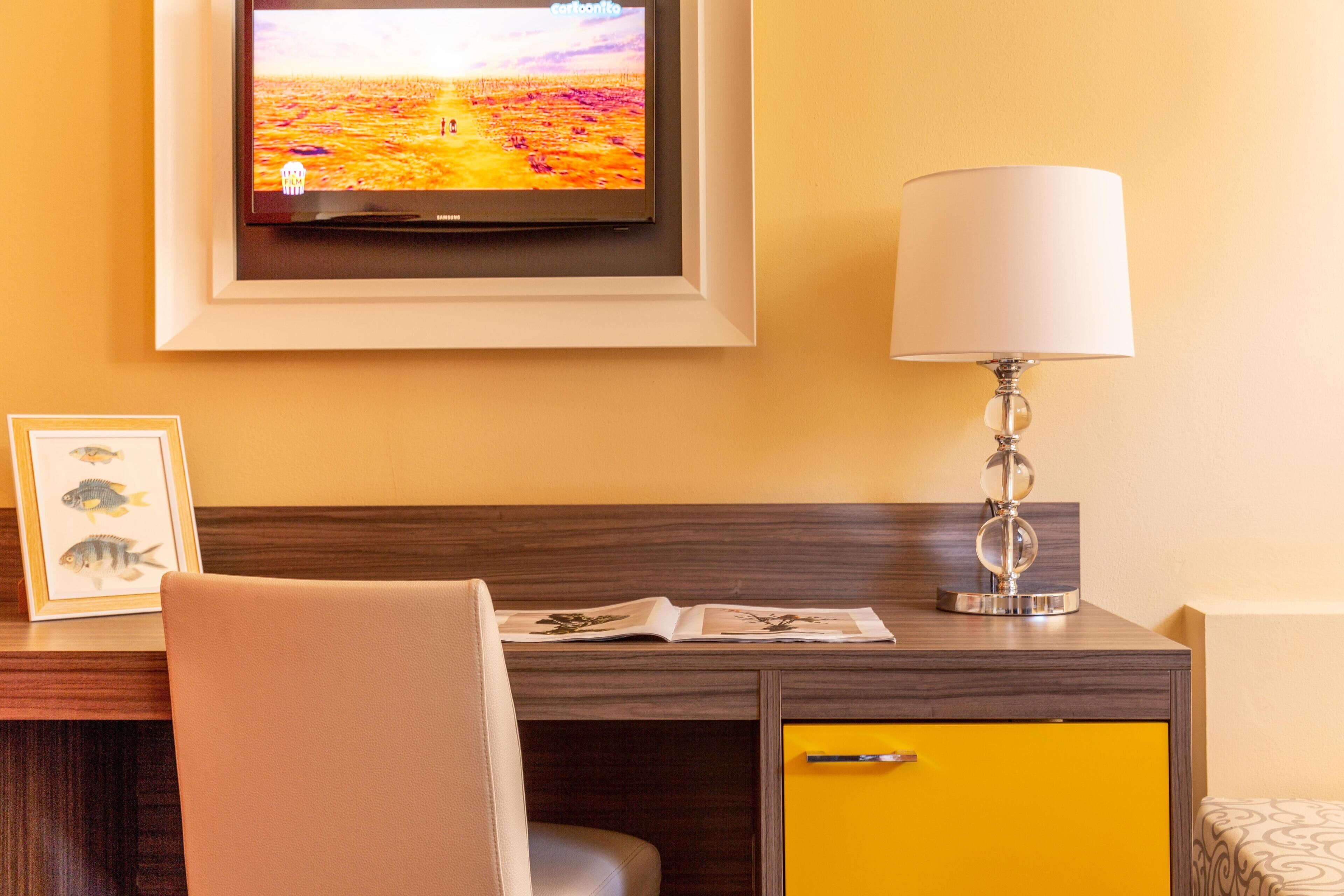 Hotellihuone, jossa on seinälle asennettu televisio, yöpöytälamppu, työpöytä tuolilla ja keltainen kaappi.