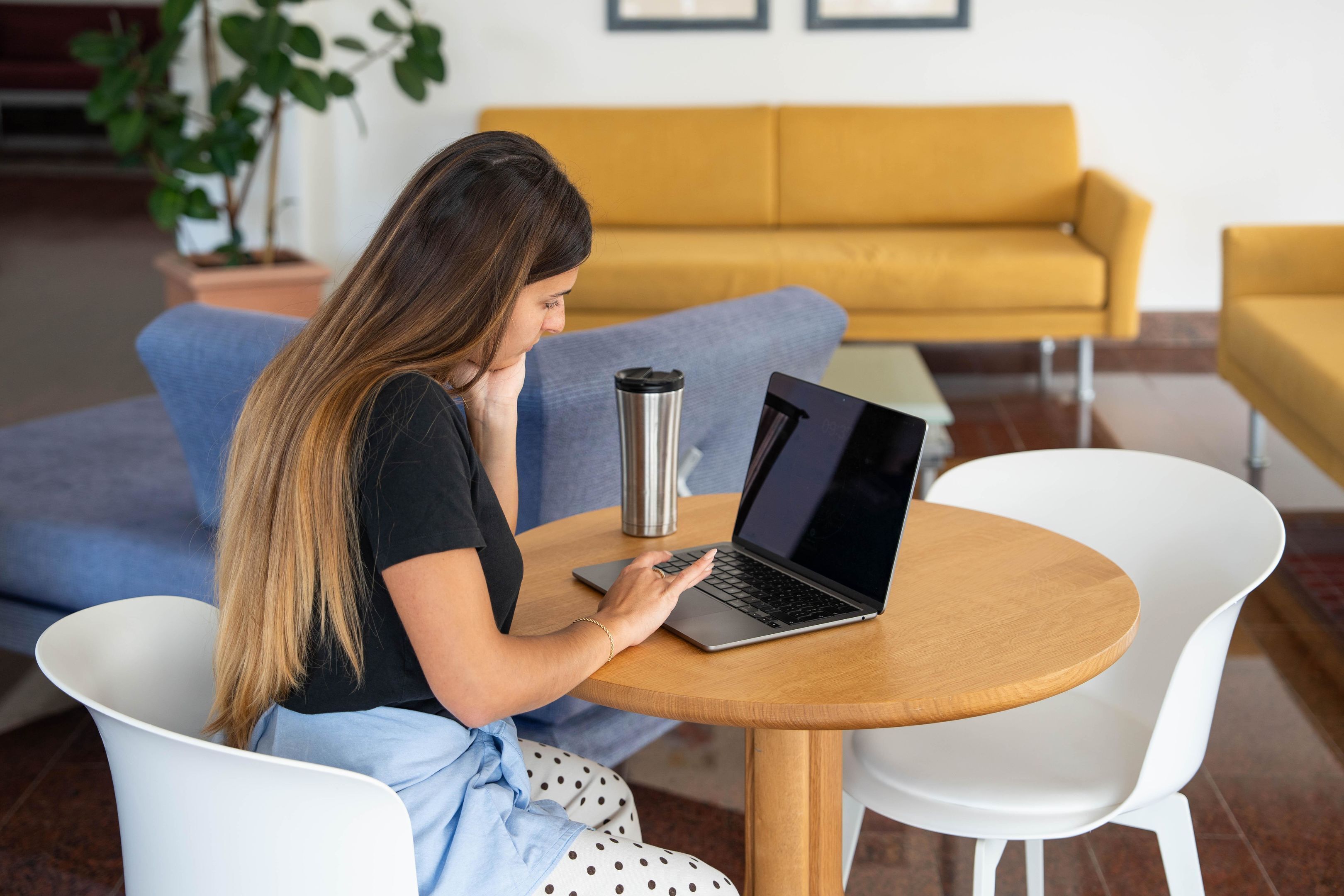 Una persona sentada frente a una mesa redonda de madera, con un ordenador portátil y un vaso de acero inoxidable, en una habitación con un sofá amarillo y sillas blancas.