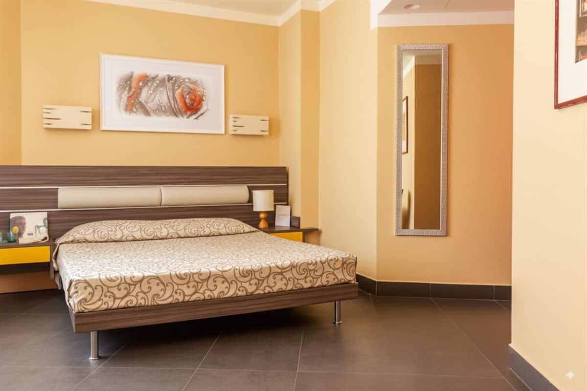 Una camera d'hotel con un letto grande, una testiera, un comodino con una lampada, un quadro incorniciato appeso alla parete e uno specchio.