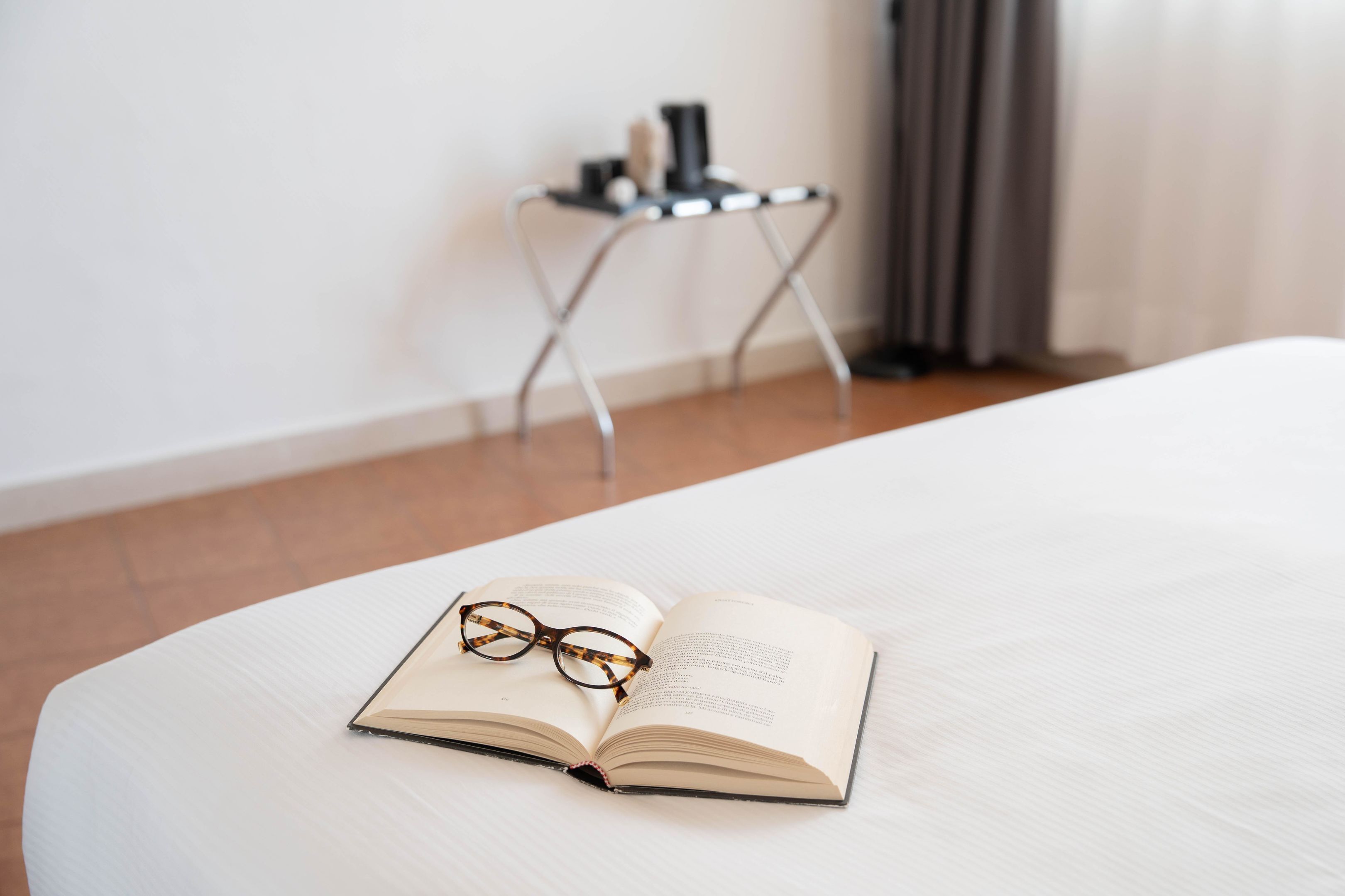 Un libro abierto sobre una cama, con gafas al lado, una mesita con una lámpara de fondo y una cortina a la derecha.