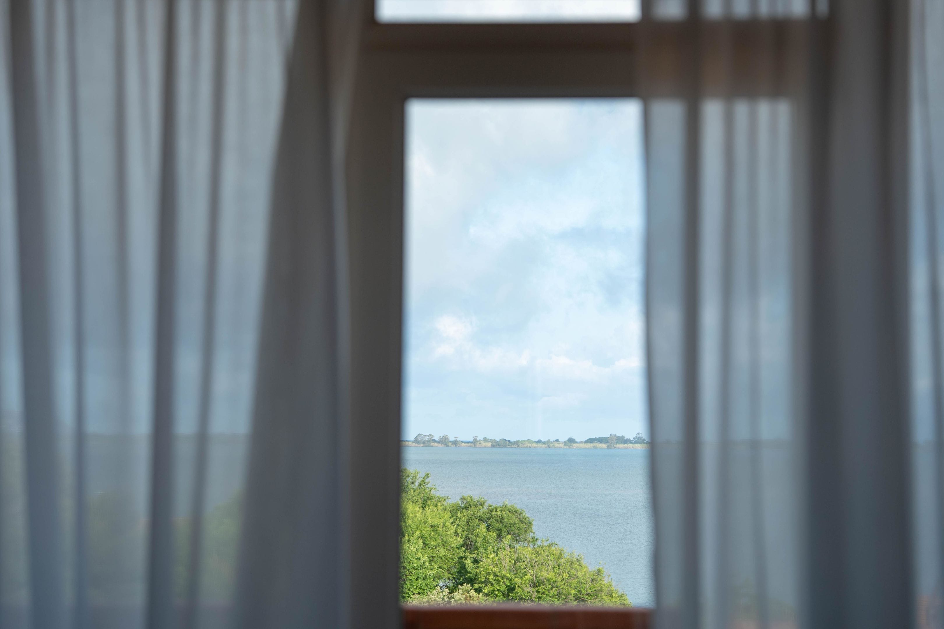 Uma janela com cortinas leves, vista para um corpo d'água e uma linha de costa distante.