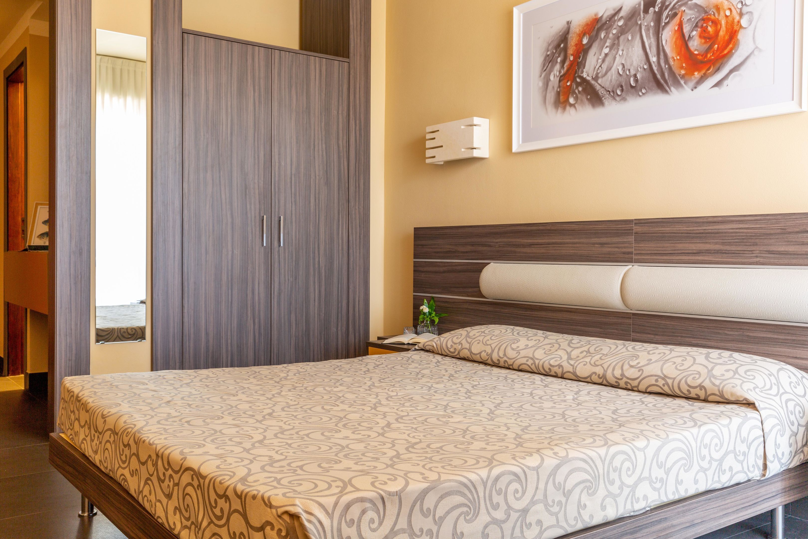 Une chambre d’hôtel comprenant un lit, une armoire, une table de chevet, une lampe fixée au mur et un tableau encadré.