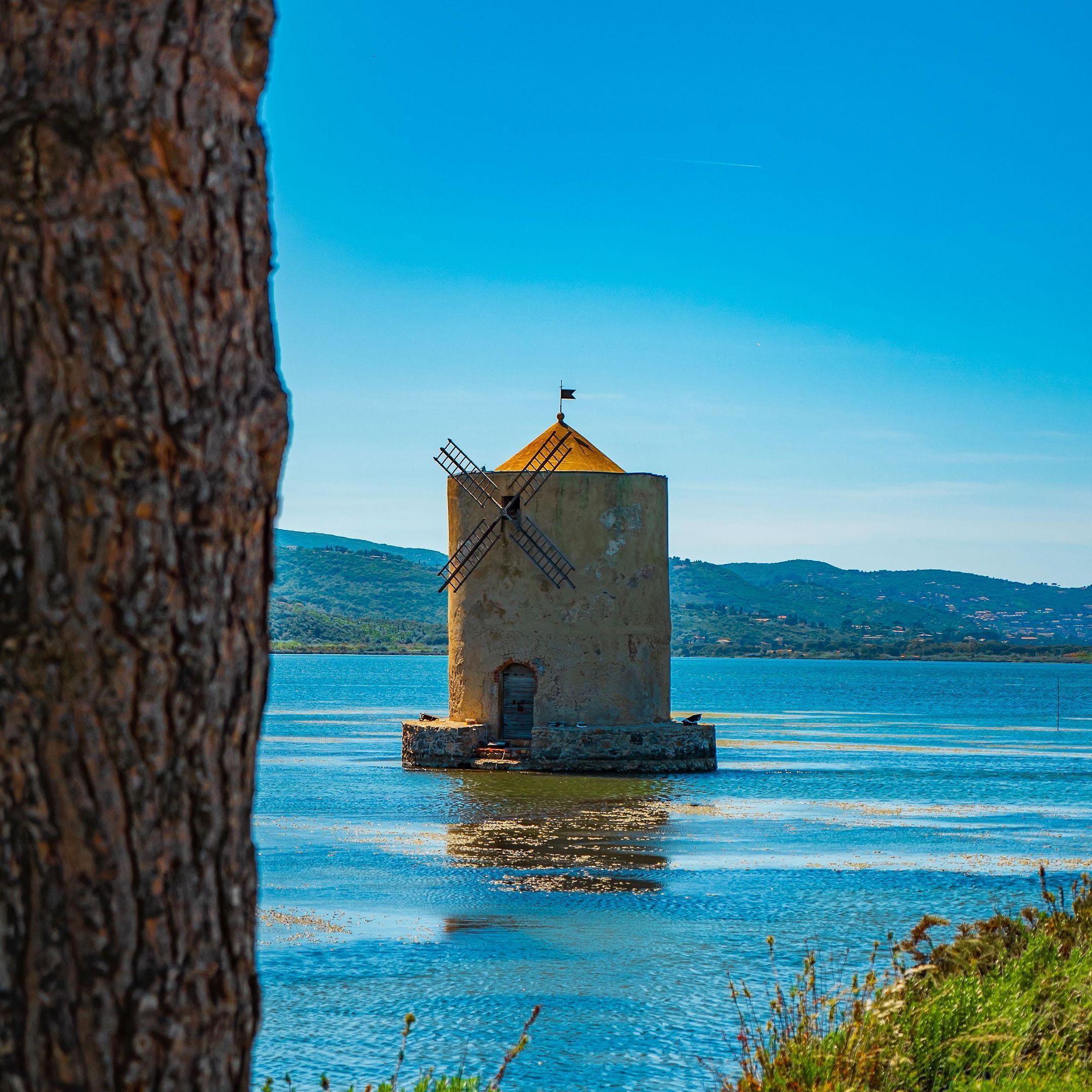 A small, isolated building with a windmill on a body of water, surrounded by hills and clear skies.