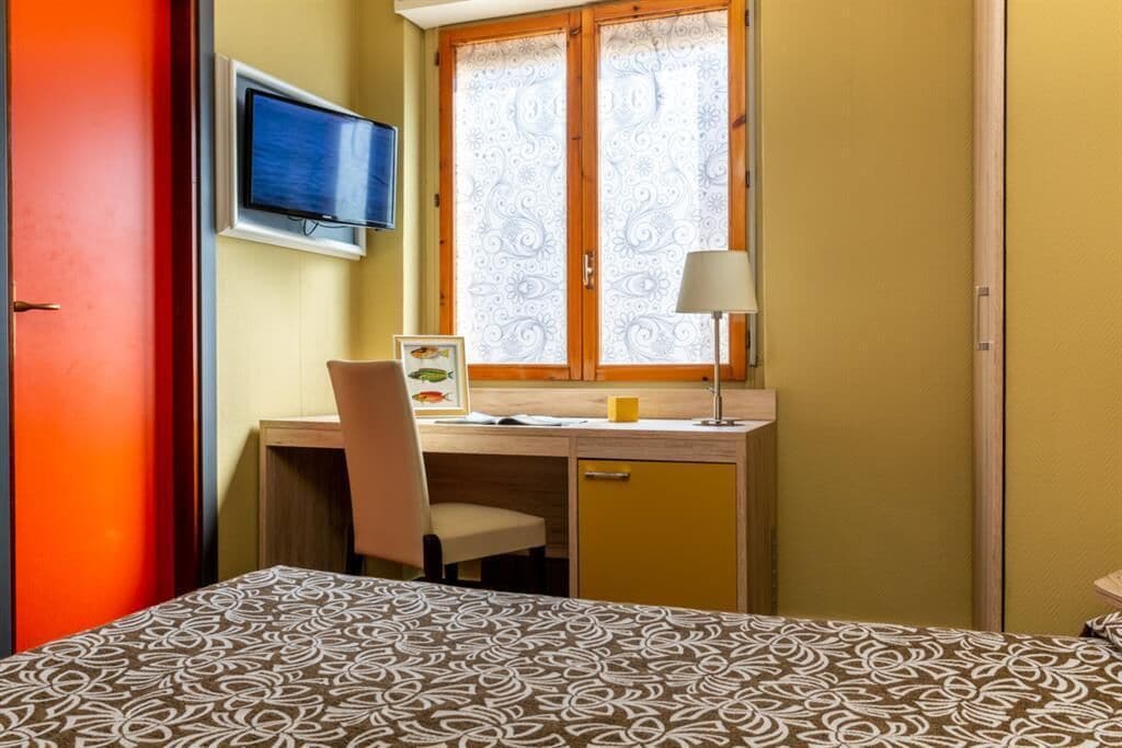Een kamer met een bed, een bureau met stoel, een televisie en een raam met gordijnen met een patroon.