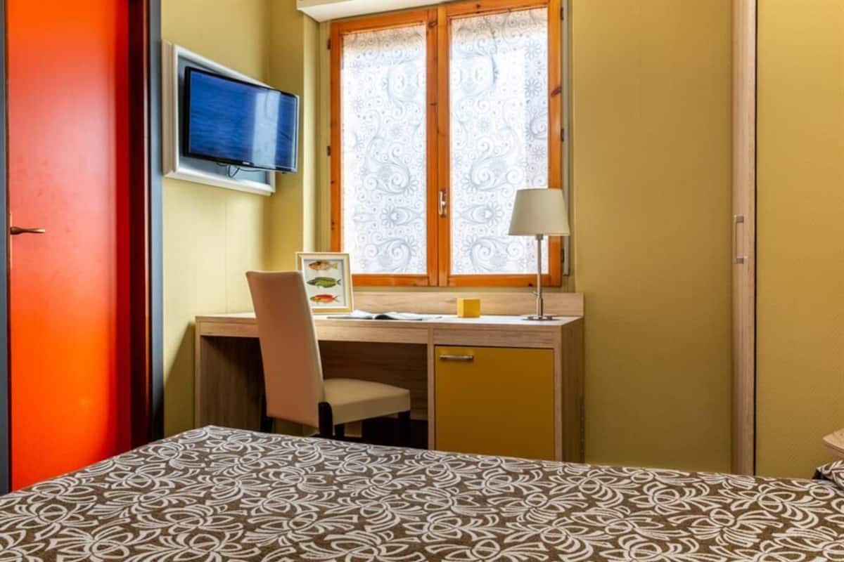 Una stanza con un letto, una scrivania con una sedia, una televisione e una finestra con tende a motivi.