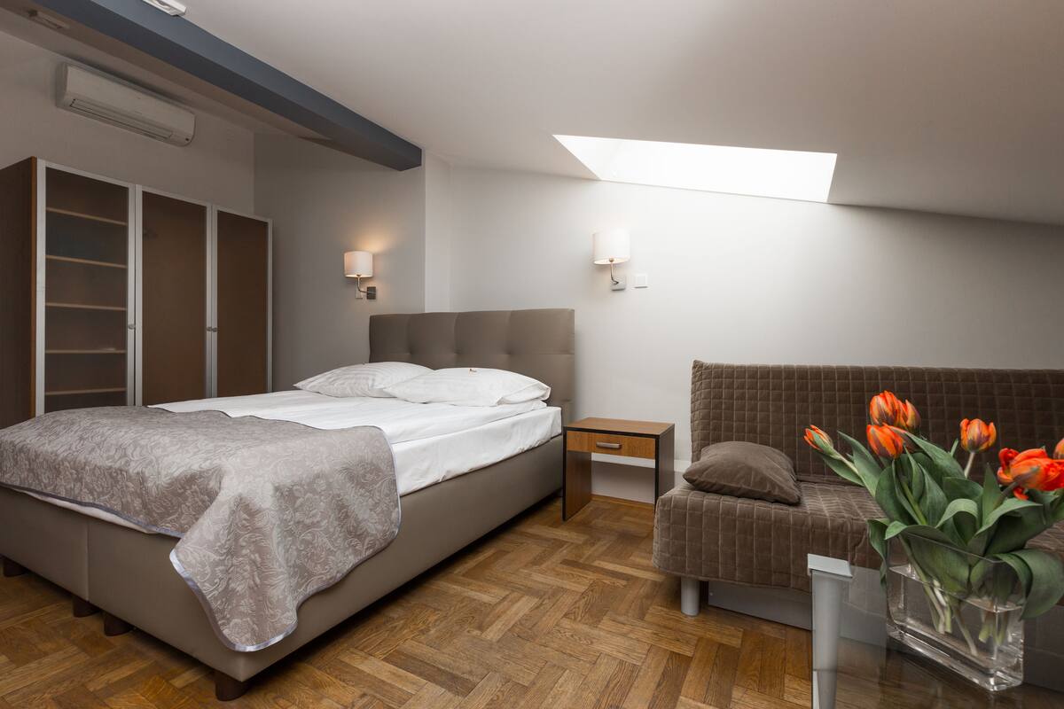 A bedroom with a bed, bedside table, nightstand, and a small seating area with a couch and a vase of flowers.