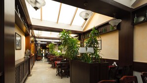 Un restaurante con techo alto, tragaluces y un macetero central con plantas verdes. Mesas y sillas dispuestas a lo largo de las paredes.